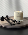 This photo shows a scented candle in a clear glass vessel on top of a decorative cement tray. There is a matte black candle snuffer and wick trimmer to the left of the candle. The try is on top of a black table with a white wall behind it.