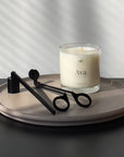 This photo shows a scented candle in a clear glass vessel on top of a decorative cement tray. There is a matte black candle snuffer and wick trimmer to the left of the candle. The try is on top of a black table with a white wall behind it.