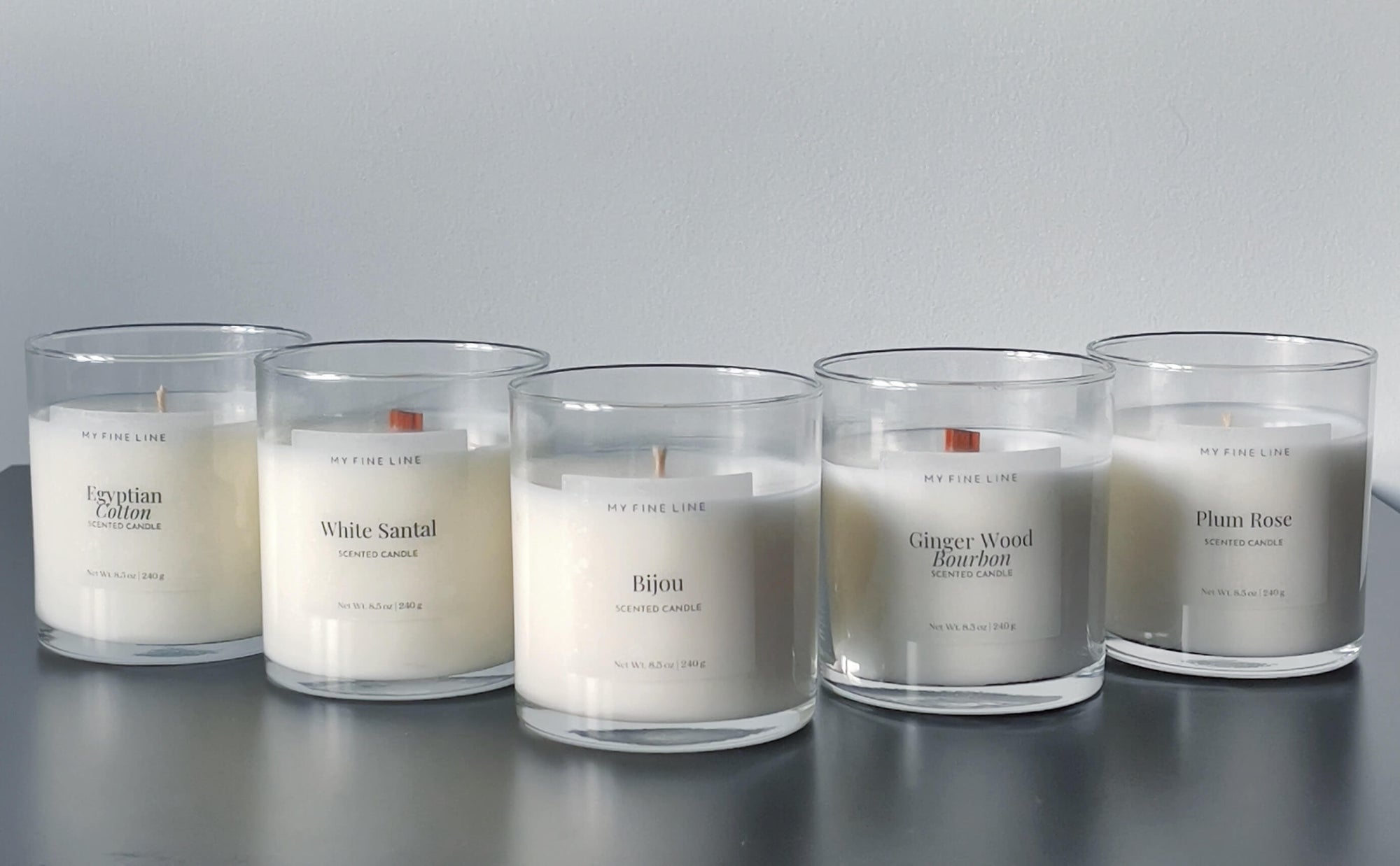 A row of five scented candles in clear glass containers, labeled from left to right: Egyptian Cotton, White Santal, Bijou, Ginger Wood Bourbon, and Plum Rose. The candles are displayed on a sleek surface against a neutral backdrop.