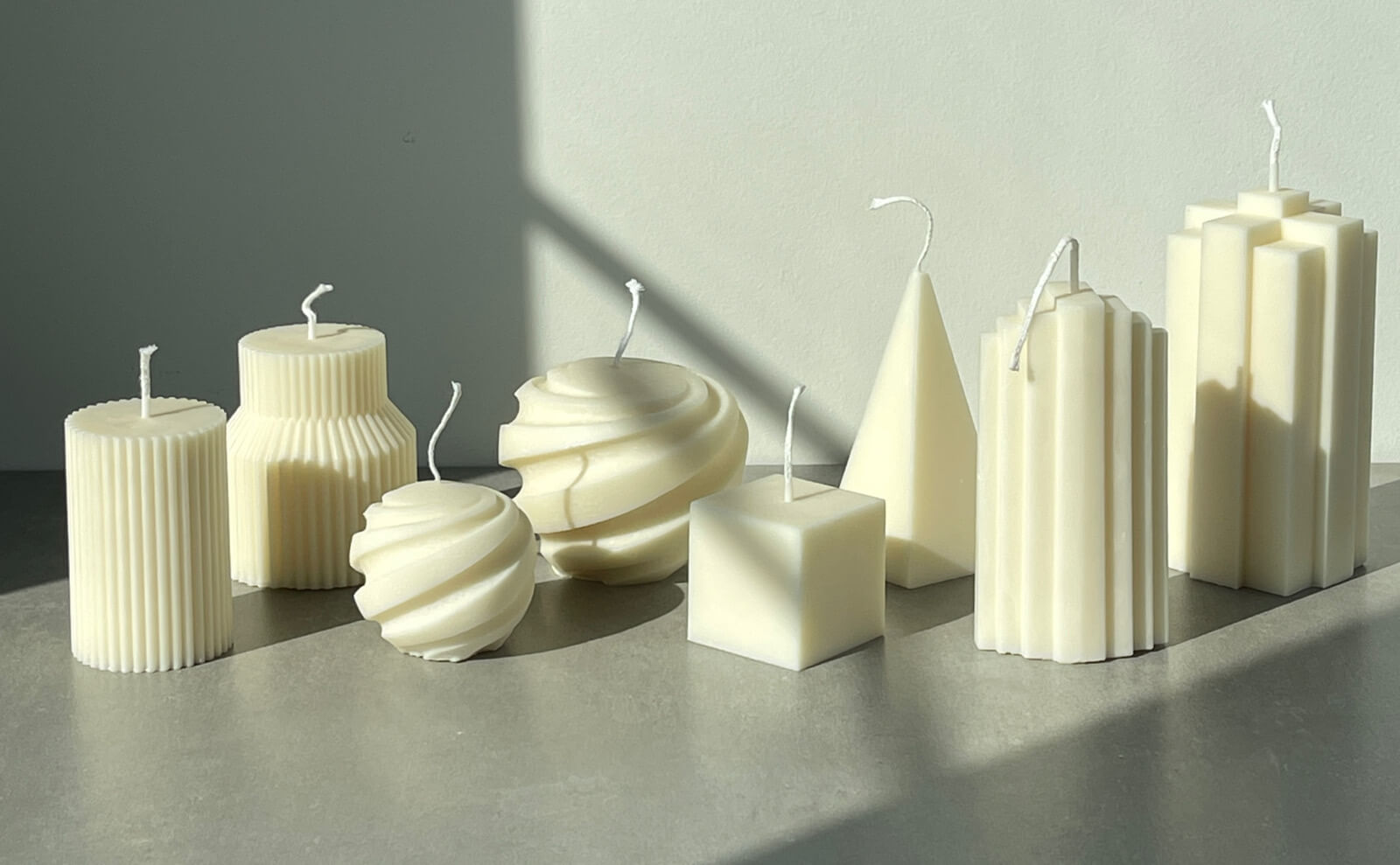 A collection of sculptural, unscented candles in various geometric shapes, arranged on a flat surface. Sunlight casts soft shadows, highlighting the minimalist design and clean lines of each candle.