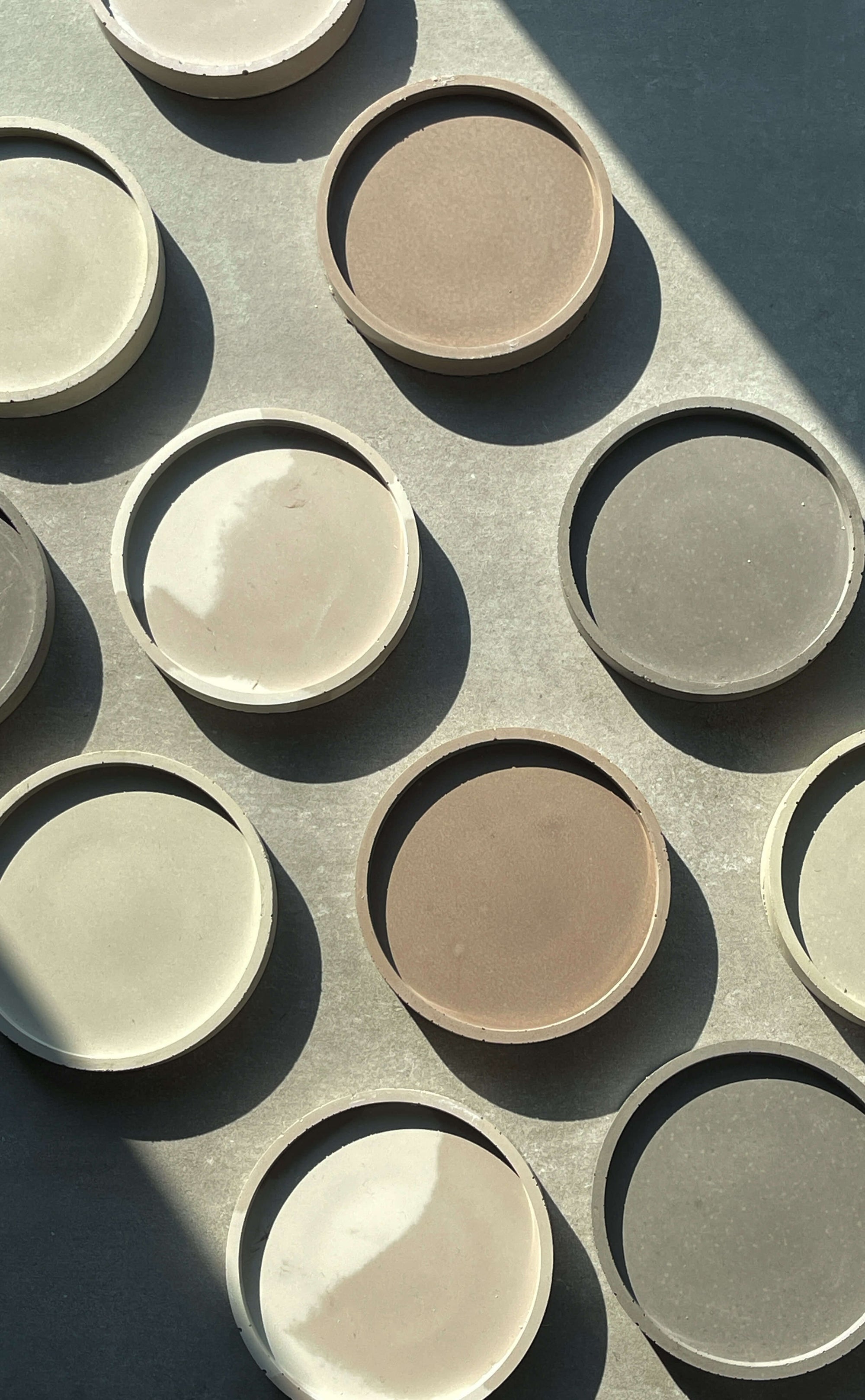 An overhead view of circular concrete trays in various neutral tones, arranged in a grid pattern. Sunlight casts soft shadows, emphasizing the textured surfaces and subtle color variations of each tray.