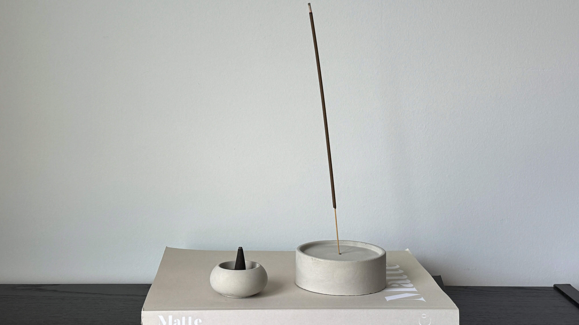 A minimalist display of incense holders on a stack of books. The larger round holder, made of concrete or similar material, holds a burning incense stick upright, while a smaller rounded holder contains a cone incense. Both items are placed on neutral-colored books, against a light gray background, reflecting a clean and modern aesthetic.