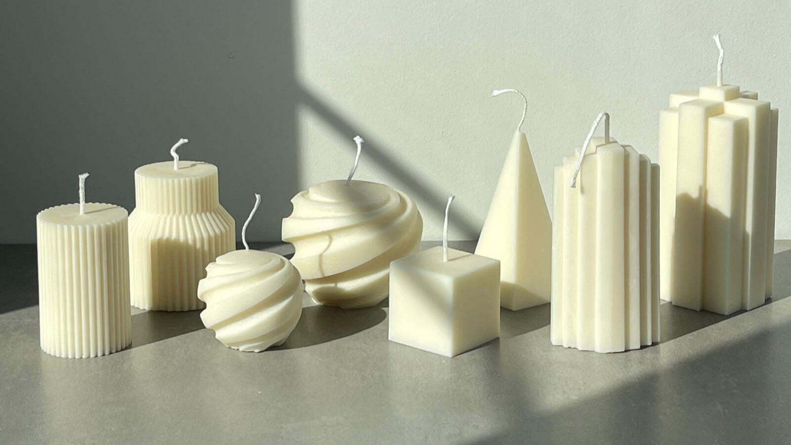 A collection of unscented, sculptural candles in various geometric shapes, including cylindrical, spiral, pyramid, and square designs. The candles are displayed on a smooth surface, illuminated by natural sunlight, casting soft shadows against a neutral gray background. The modern shapes and soft cream tones of the candles create a minimalist and elegant aesthetic.