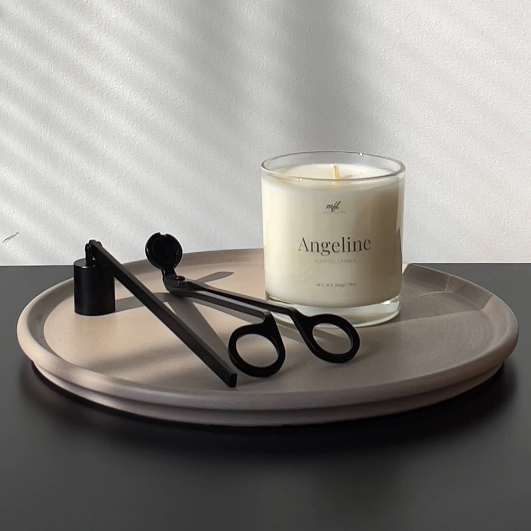 This photo shows a scented candle in a clear glass vessel on top of a decorative cement tray. There is a matte black candle snuffer and wick trimmer to the left of the candle. The try is on top of a black table with a white wall behind it.