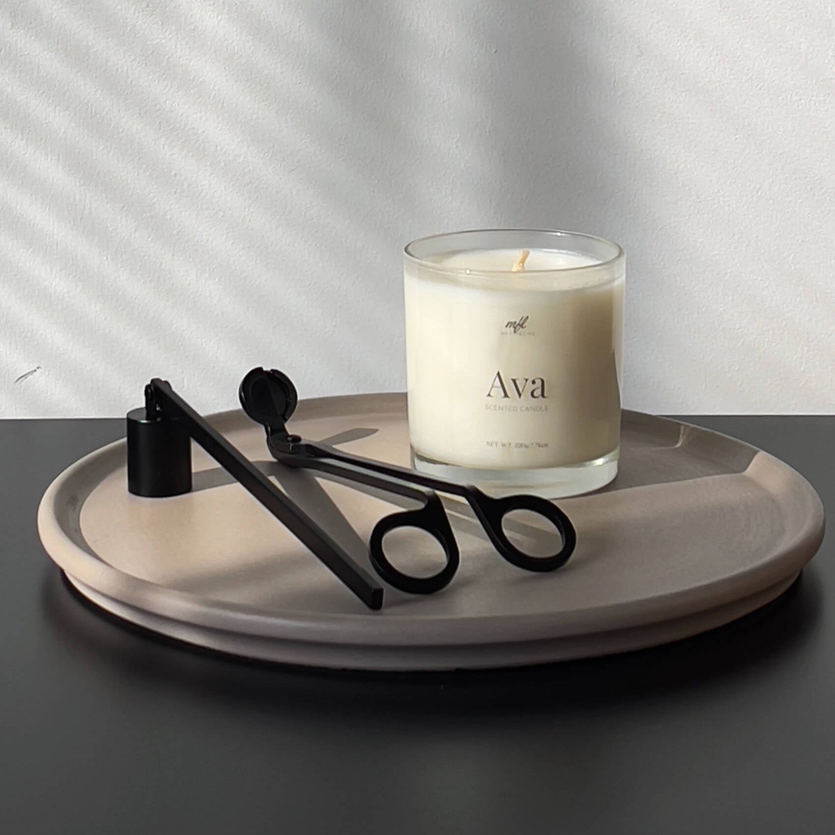 This photo shows a scented candle in a clear glass vessel on top of a decorative cement tray. There is a matte black candle snuffer and wick trimmer to the left of the candle. The try is on top of a black table with a white wall behind it.